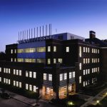 Four-story rectangular red brick building at dusk with all the lights on. There is a row of outdoor lights on the right-side of the building along the sidewalk and three cars parked along the left-side of the building.