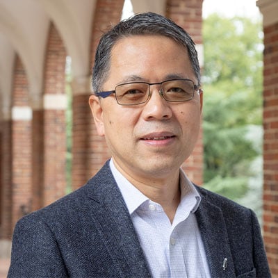 Headshot of Hai-Quan Mao. He has salt and pepper short hair and light to medium skin tone. He is wearing square, black framed glasses, a light blue button down dress shirt, and a gray tweed suit jacket. He is standing outside under a pathway with brick arches in the background.
