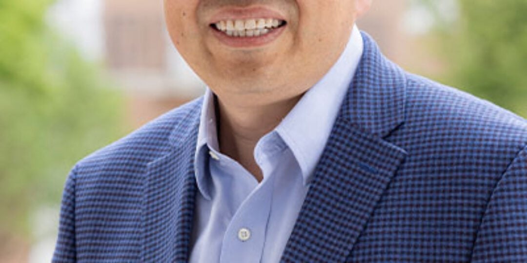Headshot of Honggang Cui. He has short salt and pepper hair and light to medium tone skin. He is wearing square framed glasses, a blue button down dress shirt, and a blue and black plaid suite jacket. He is standing outside with a redbrick building and green trees in the background.