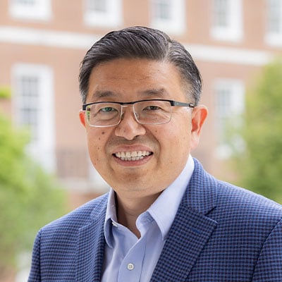 Headshot of Honggang Cui. He has short salt and pepper hair and light to medium tone skin. He is wearing square framed glasses, a blue button down dress shirt, and a blue and black plaid suite jacket. He is standing outside with a redbrick building and green trees in the background.