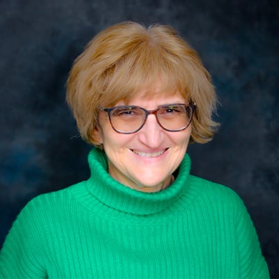 Headshot of Kalina Hristova. She is wearing a green turtleneck sweater and black square rimmed glasses. She has light skin tone, cheekbone length blonde hair with bangs, and dark color eyes.