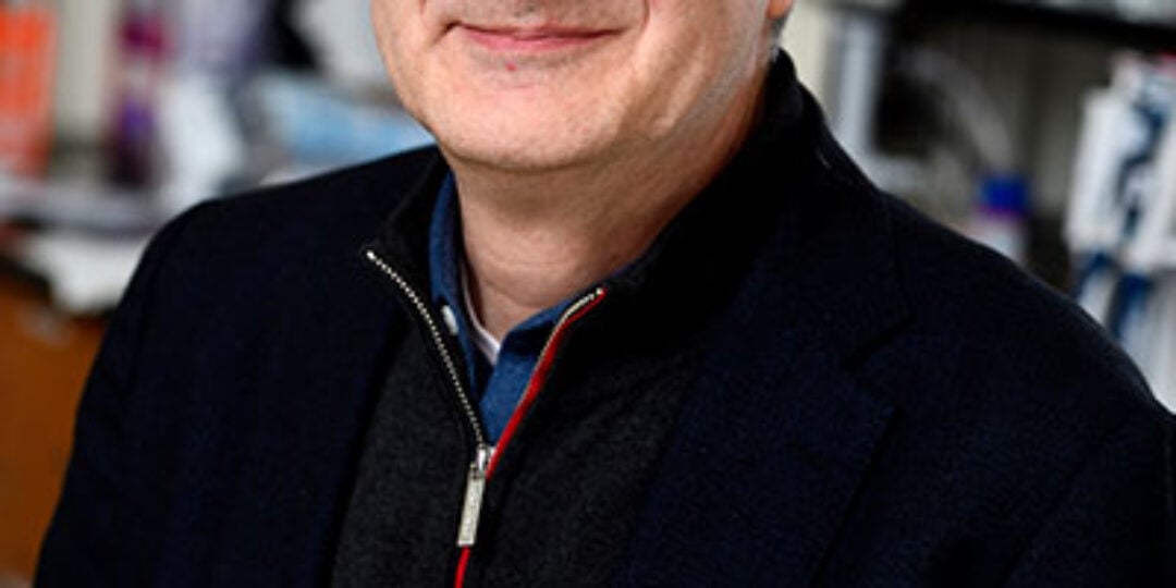 Headshot of Denis Wirtz. He has short salt and pepper hair and light skin tone. He is wearing black square framed glasses, a black quarter zip pull over and a blue button down dress shirt. In the background is a laboratory.