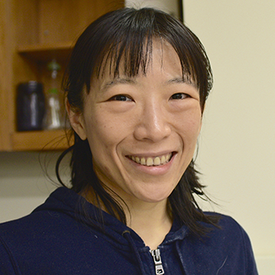Headshot of Yun Chen. She is standing in front of a laboratory cabinet on the wall is wearing a blue zip up sweater.