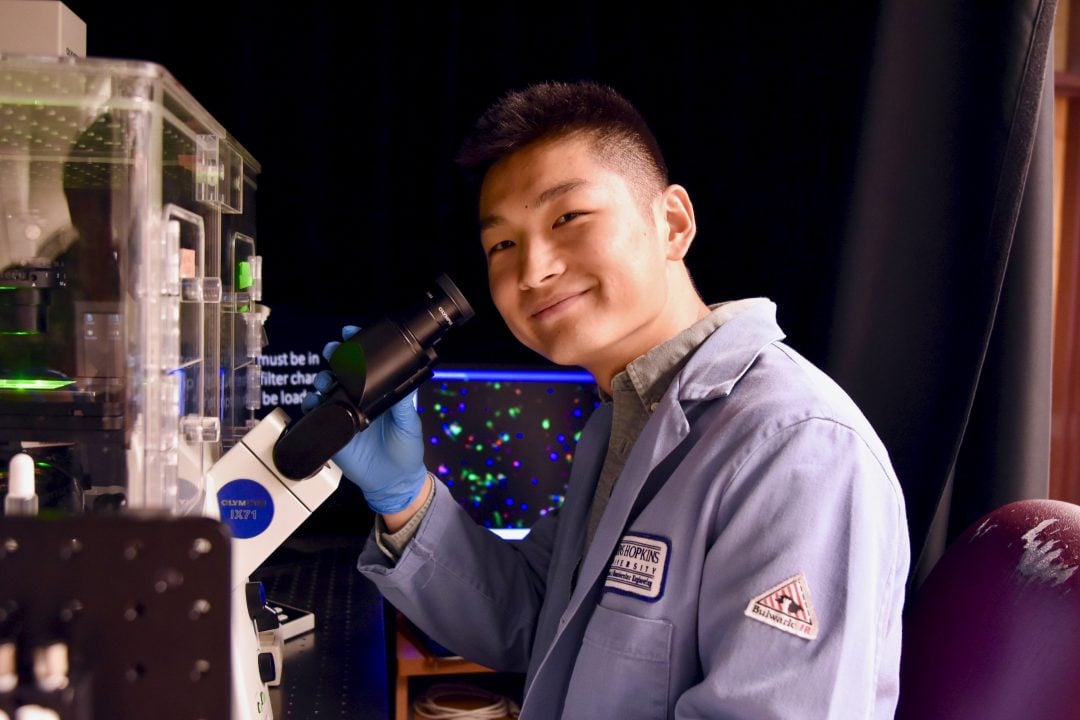 Man in blue lab coat looking through an electron microscope.