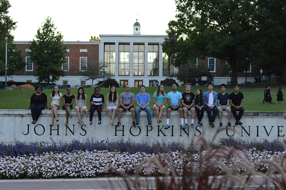 16 students sitting on a marble wall with the Johns Hopkins University name on it. In the background is a red brick and glass library.