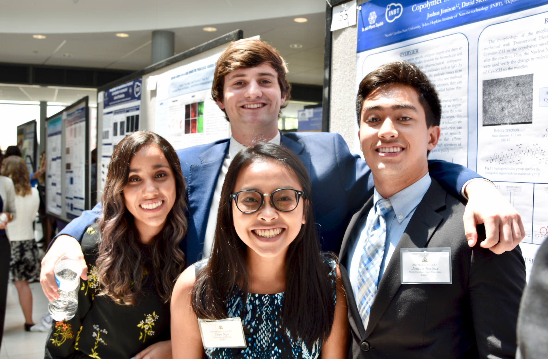 Group photo of four students in formal clothes at the 2018 CARES Symposium. In the background is a row of poster boards with research posters.