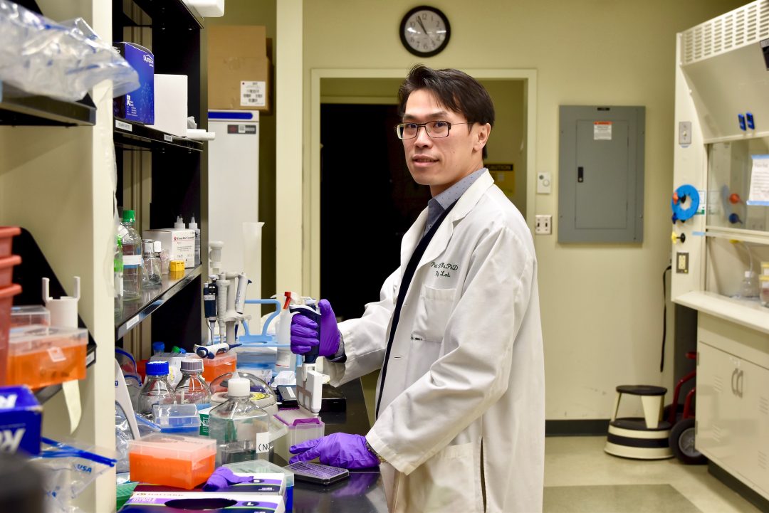 Researcher in a white lab coat standing in front of a black-top lab bench. He is pipetting a liquid material with other lab supplies around him.