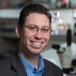 Headshot of Jordan Green. He is wearing a dark blazer over a button down blue dress shirt. He is standing in a laboratory with a blurred background. He has short dark hair, light skin, brown eyes, and is wearing square framed glasses.