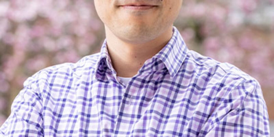 Headshot of Sangmoo Jeong. He has black hair, brown eyes, and light skin tone. He is wearing round black glasses and a pink, blue, and white plaid button down dress shirt. He is standing outside with cherry blossom trees blooming in the background.