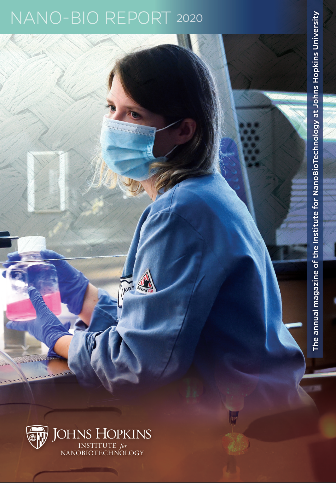 Magazine cover image. Woman in blue lab coat and blue mask working in a research lab. She is under a fume hood and holding a bottle with pink fluid.