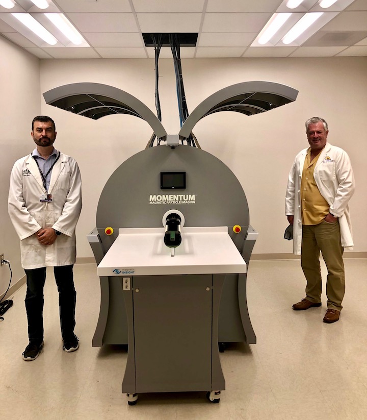 Adnan Bibic is standing on the left in a white lab coat and Jeff Bulte is standing on the right in a white lab coat. In between them is a large cylidrical machine with doors open on either side and a long table in front of it.