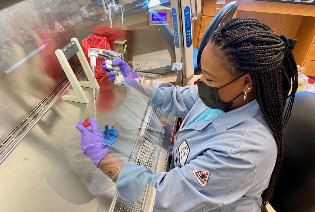 A young African American women in a blue lab coat, purple gloves, and black face mask is pipetting a liquid under a fume hood in a laboratory.