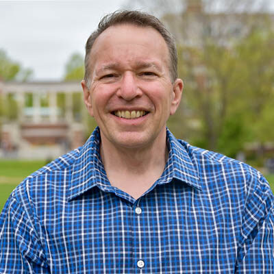 Headshot of Tim Allgire. He has light skin and short hair and is wearing a blue and white plaid pattern dress shirt. He is standing outside with a brick and marble building in the background and a tree starting to bloom in the spring.