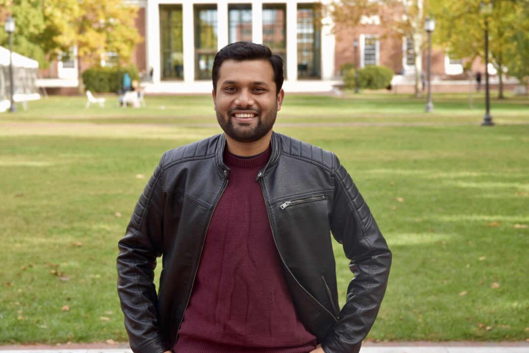 Photo of Ikbal Choudhury wearing a maroon sweater under a black leather jacket. He is standing in front of an long grass field with a brick and white building in the background.