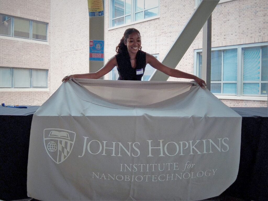 Woman laying out a gray table cloth with the words Johns Hopkins Institute for NanoBioTechnology over a rectangular table with black table clothes.