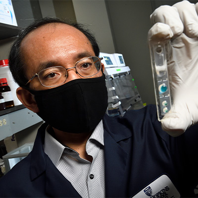 Research Jeff Wang stands in a laboratory holding and looking at a microfluidic device. He is wearing glasses, a black face mask, a dark blue lab coat and white nitrile gloves.