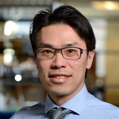 Headshot of Pei-Hsun Wu. He is wearing a blue stripped collared shirt with a blue and gray diamond pattern tie. He is standing in front of a blurred laboratory setting. He has short black hair, dark eyes and is wearing black rectangular glasses.