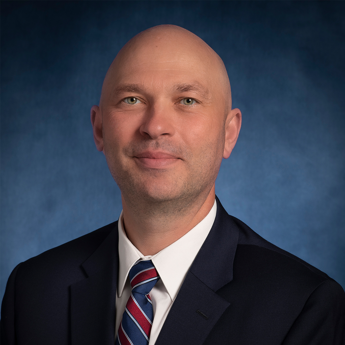 Headshot of Florin Selaru. He is without hair and has green eyes. He is wearing a dark blue blazer over a white button down dress shirt and blue and red stripe tie.