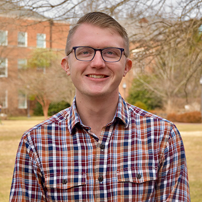 Headshot of Johnny Moseman. He has blonde hair, blue eyes, and light-colored skin. He is wearing dark, square rimmed glasses and a plain button down collared shirt. He is standing in front of a large lawn and brick building in the background.