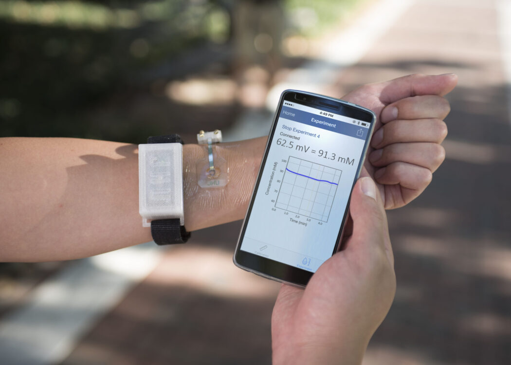 Picture of left arm with a skin sensor on the wrist. In the right hand is a mobile phone with a table on it indicating health information.