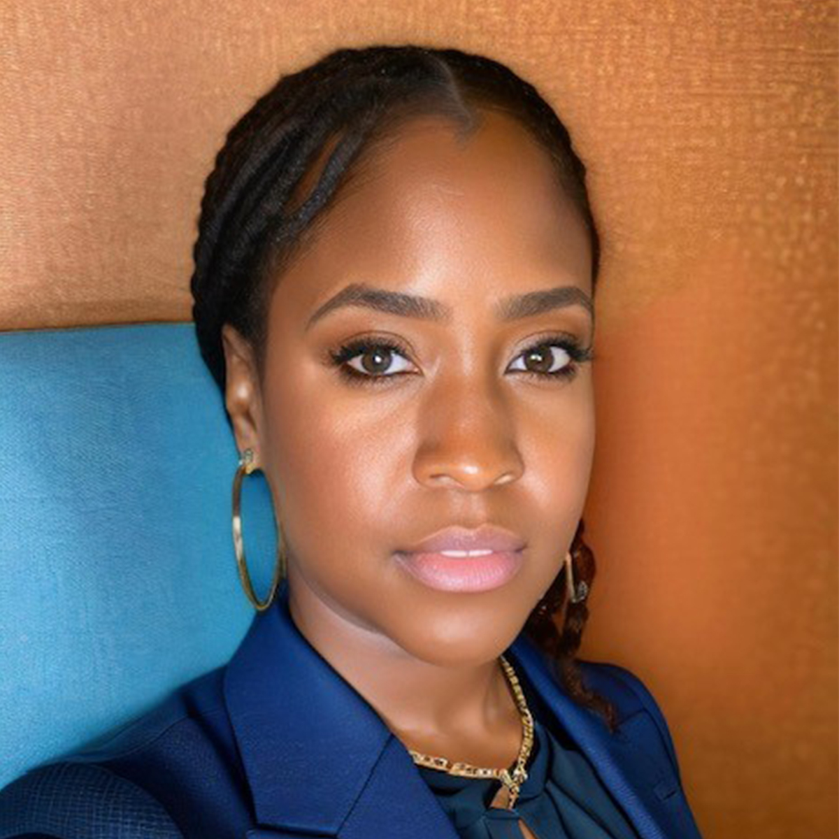 Headshot of Ashely Moore. She brown eyes, dark skin, and long hair. She is wearing a dark blue blazer over a dark blue button down blouse. She is also wearing gold hoop earrings and gold braided necklace. She is standing in front of a terra cotta and blue background.