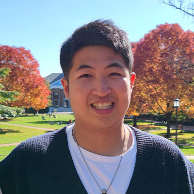 Headshot of Yining Zhu. He has short dark hair and brown almond shaped eyes. He is wearing a black v-neck sweater over a white crew neck t-shirt. He is standing outside with fall colored trees in the background on a campus quad.