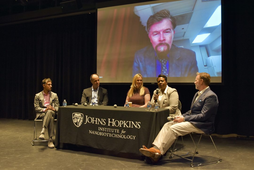 Panel discussion with five people sitting at a rectangular table and one virtual speaker projected on a drop down screen. They are in a theater.