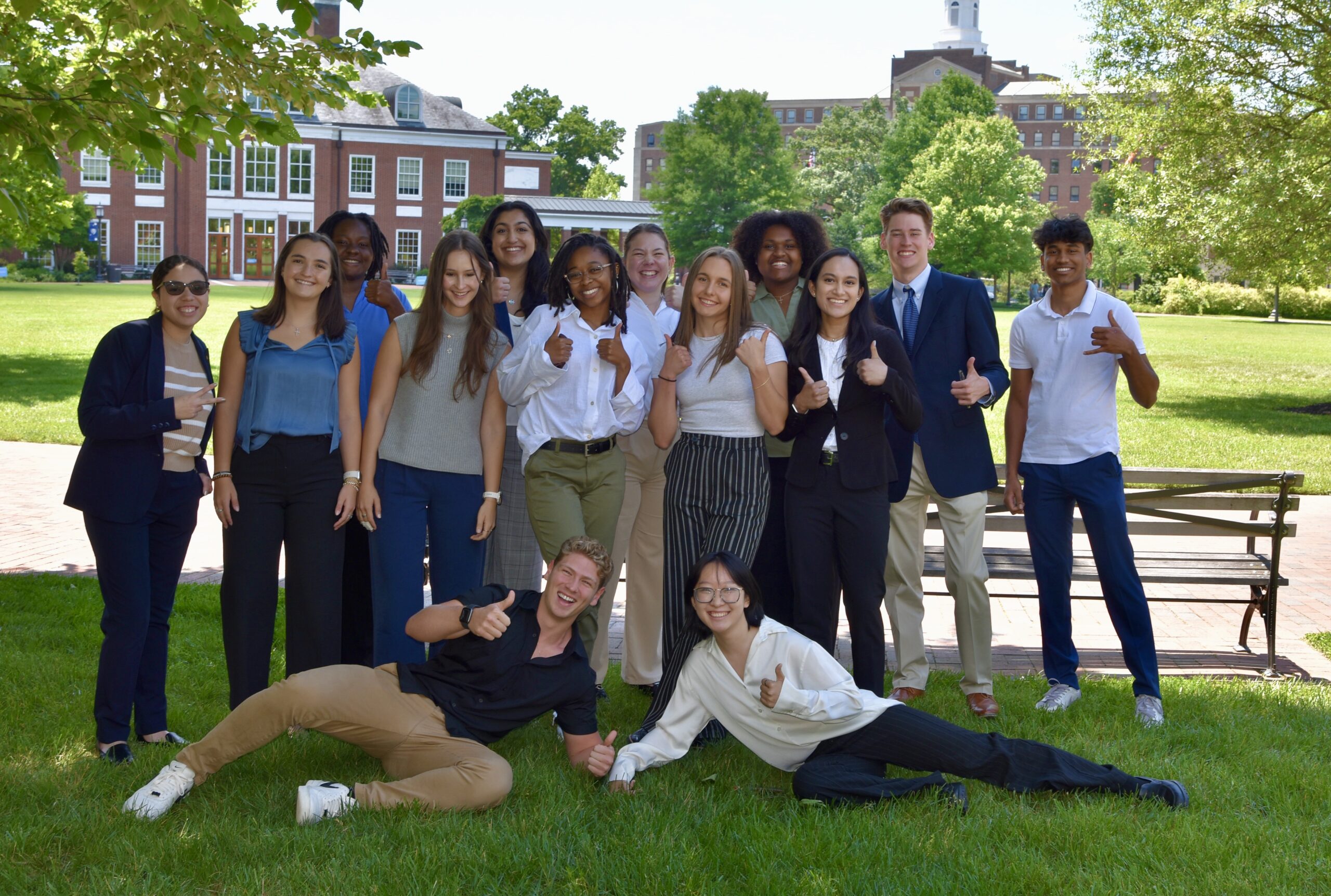 Group photo of 14 students in business casual clothes smiling at camera. They are under the shade of a tree on a campus quad. In the background are red brick buildings and an open field.