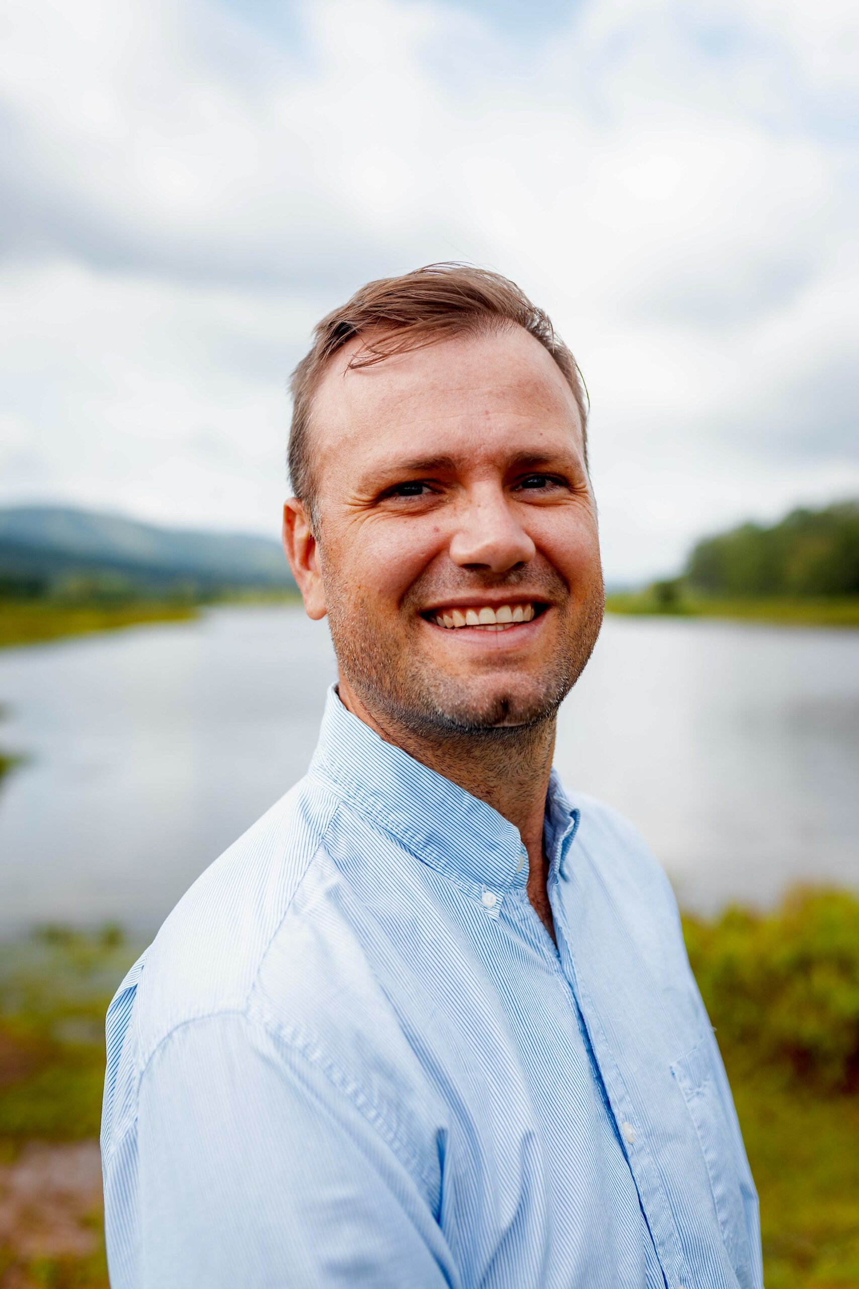 Headshot of Luke Thorstenson. He has fair skin and short brown hair. He is wearing a blue stripped button down dress shirt and standing outside in front of a lake.