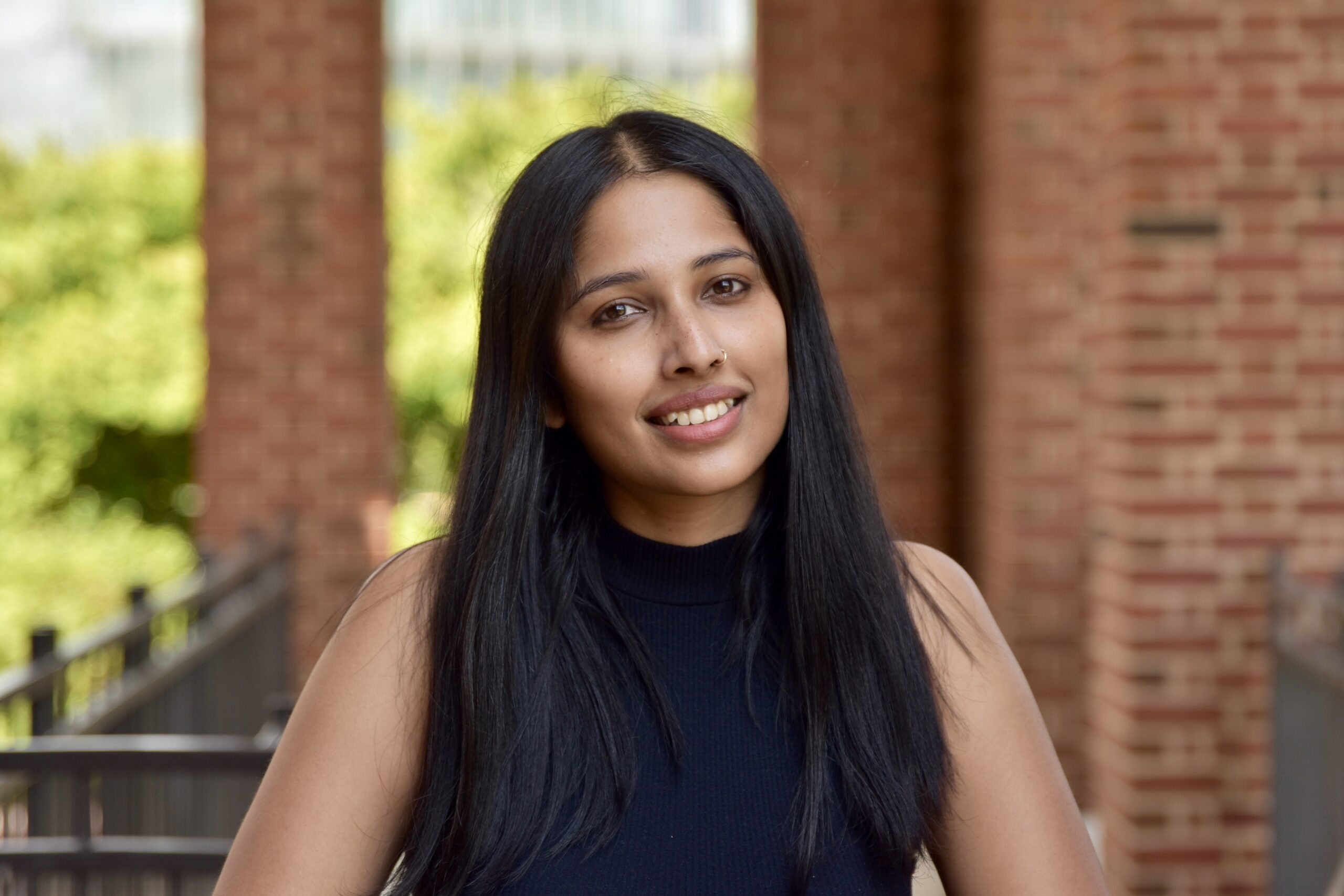 Headshot of Tanaya Roy. She has brown eyes, long straight black hair, and medium skin tone. She is wearing a black sleeveless turtle neck and standing in front of red brick columns.