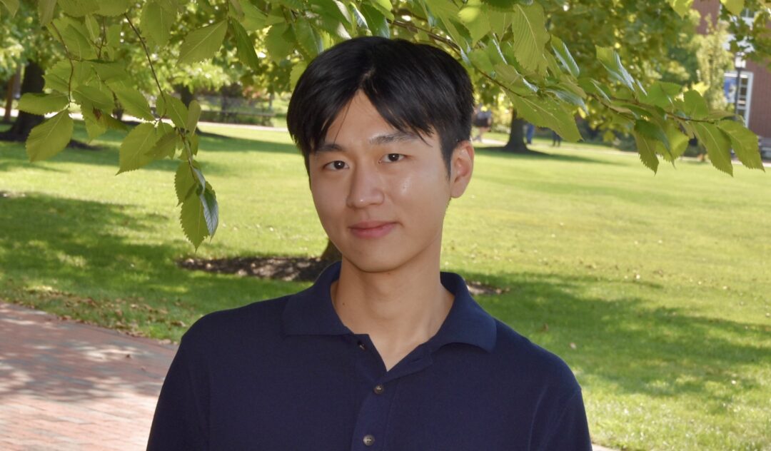 Headshot of Heonjoon Lee. He has dark eyes and short black hair with bangs. He is wearing a dark blue polo shirt and standing under a tree in front of a campus quad.