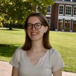 Headshot of Emily Help. She has light skin, blue eyes, and light brown hair. She is wearing round black frame glasses, small hoop earrings, and a v-neck beige shirt. She is standing in front of a grass field with a tree and red brick building in the background.