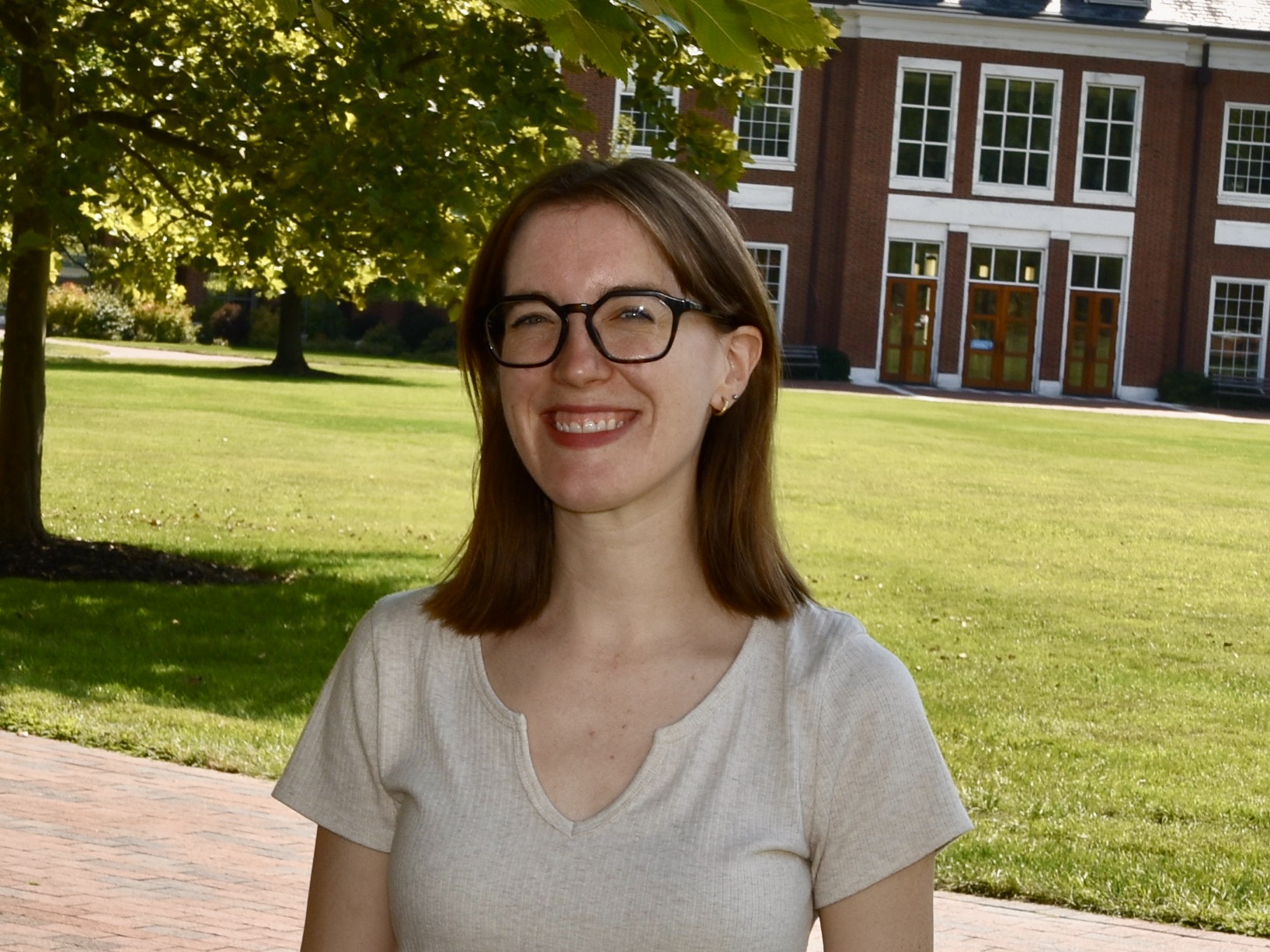 Headshot of Emily Help. She has light skin, blue eyes, and light brown hair. She is wearing round black frame glasses, small hoop earrings, and a v-neck beige shirt. She is standing in front of a grass field with a tree and red brick building in the background.