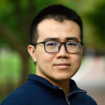 Headshot of Dingchang Lin. He is wearing a dark blue quarter zip pull over and black square glasses. He has short black hair, brown eyes, and is standing outside in front of a tree with a blurred background.