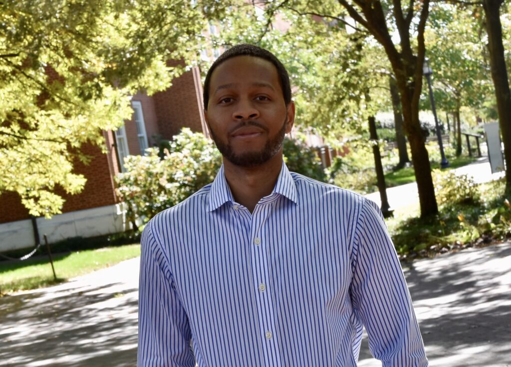 Headshot of Eban Hanna. He has a medium-dark skin tone, brown eyes, short dark hair, and a short beard and mustache. He is wering a button down dress shirt in white and blue vertical stripes. He is standing outside under trees with a red brick building in the background.