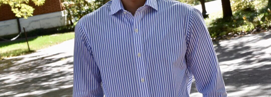 Headshot of Eban Hanna. He has a medium-dark skin tone, brown eyes, short dark hair, and a short beard and mustache. He is wering a button down dress shirt in white and blue vertical stripes. He is standing outside under trees with a red brick building in the background.