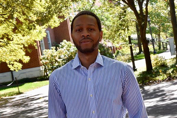 Headshot of Eban Hanna. He has a medium-dark skin tone, brown eyes, short dark hair, and a short beard and mustache. He is wering a button down dress shirt in white and blue vertical stripes. He is standing outside under trees with a red brick building in the background.