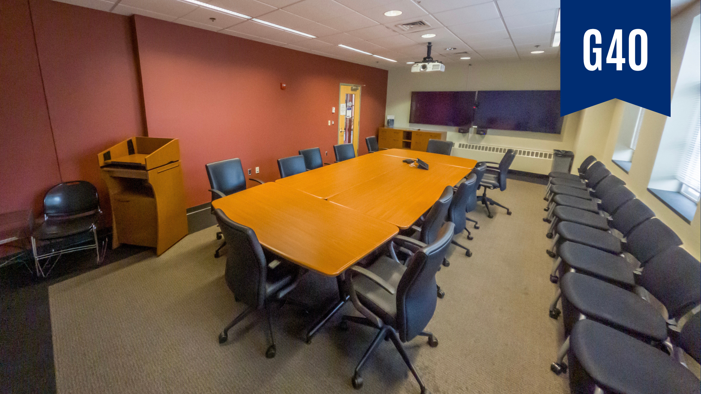 Rectangular room with white board at one end and two large monitors for Zoom meetings at the other. In the center is a rectangular table surrounded by leather computer chairs. Along the wall is a row of chairs.