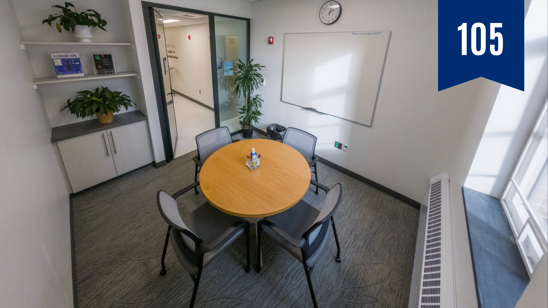 Picture of a small conference room. In the center is a small round table surrounded by four chairs on caster wheels. There are plants in the far corner of the room and on shelves to the left.