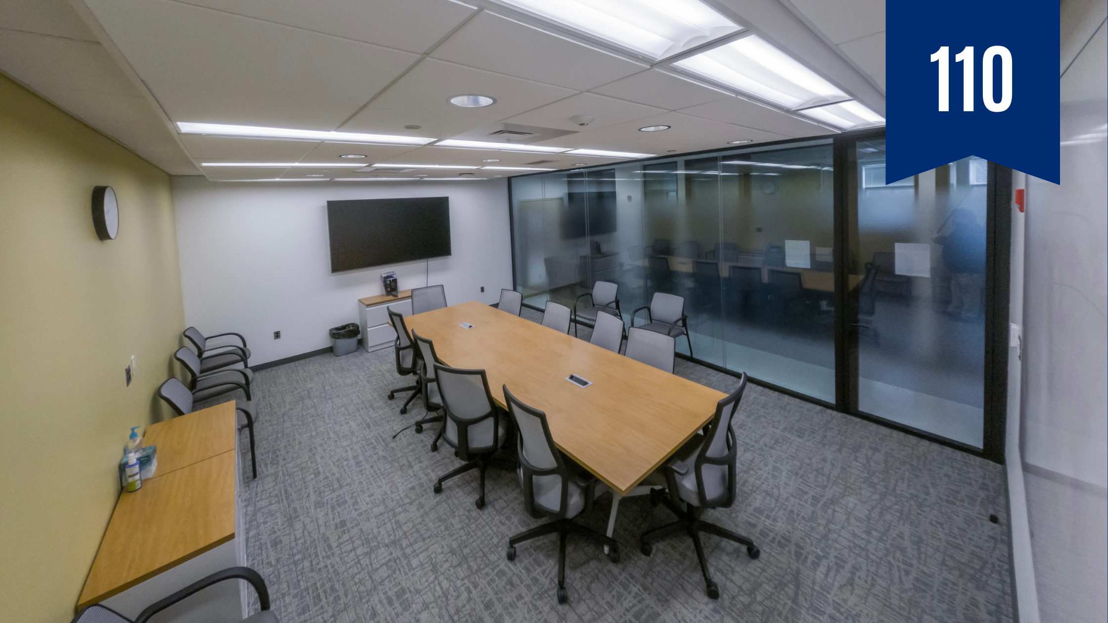 Conference room with large rectangular table in the center with ten chairs around it. The wall in the background is yellow and there is a large television on the opposite wall.