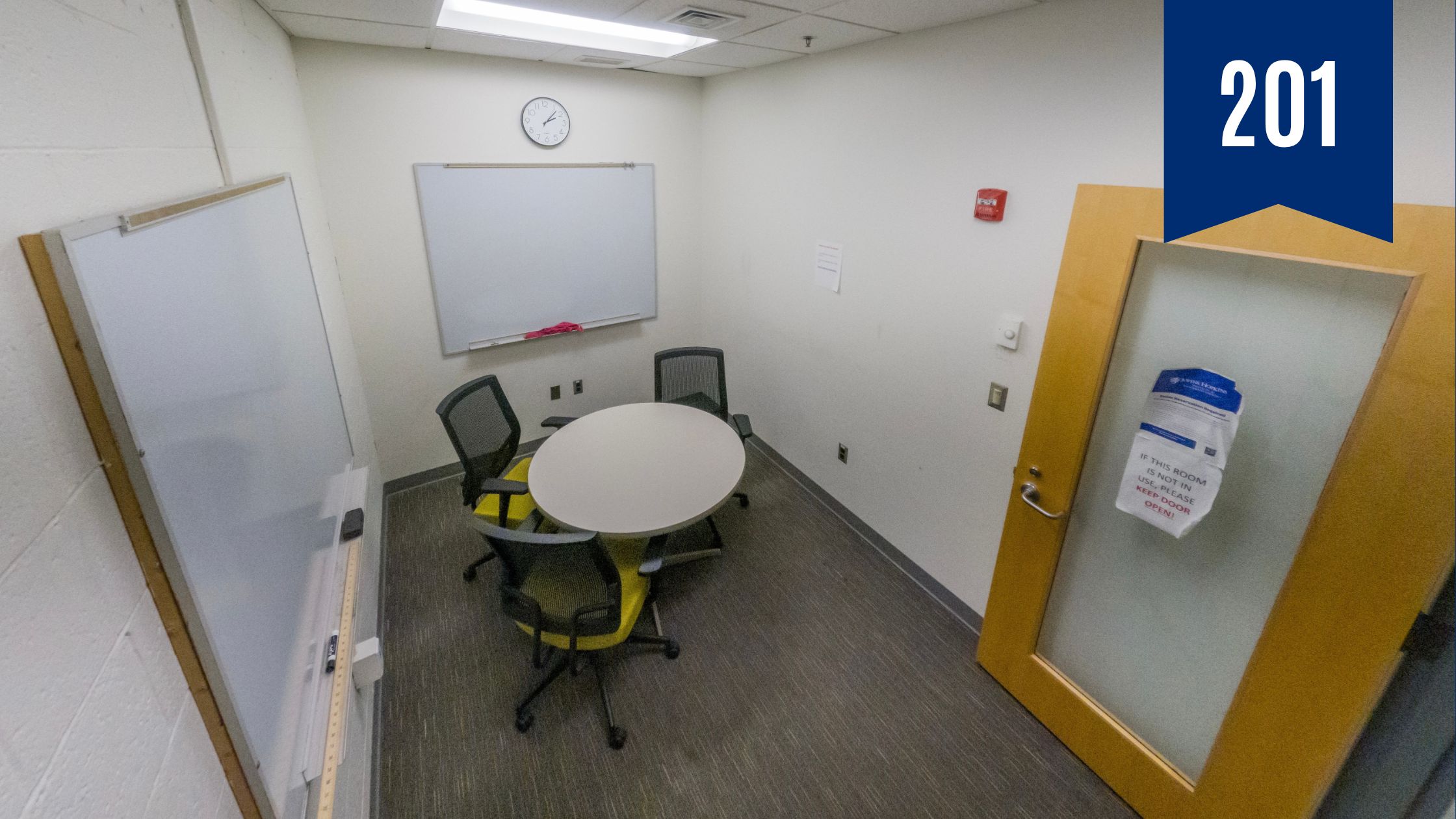 Small conference room with round table in the center and three chairs around it. Two walls have white boards and there is one blue chair in the corner
