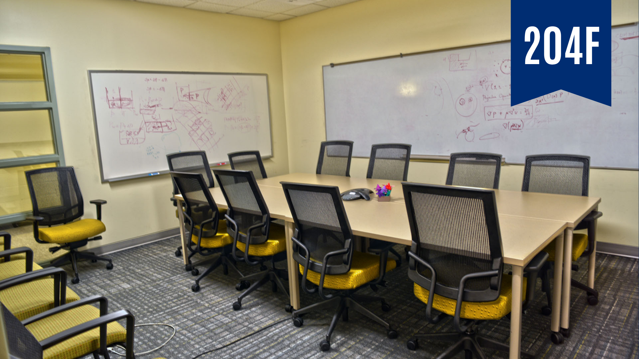 Rectangular room with two white boards. In the center is a rectangular table surrounded by 10 computer chairs. Along the wall is a row of four computer chairs.