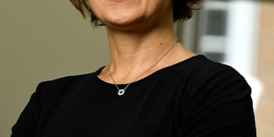 Headshot of Efrosini (Efie) Kokkoli in an office. She is wearing a black scoop neck top and silver necklace with a circle pendant. She has short, wavy brown hair, brown eyes, and medium-light skin tone.