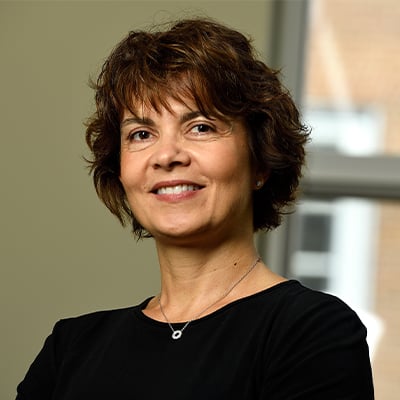 Headshot of Efrosini (Efie) Kokkoli in an office. She is wearing a black scoop neck top and silver necklace with a circle pendant. She has short, wavy brown hair, brown eyes, and medium-light skin tone.