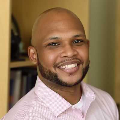 Headshot of Jude Phillip. He has brown eyes, medium-dark skin tone, no hair, and a dark mustache and beard. He is wearing a red and white plaid dress shirt. He is in an office with a bookshelf in the background.
