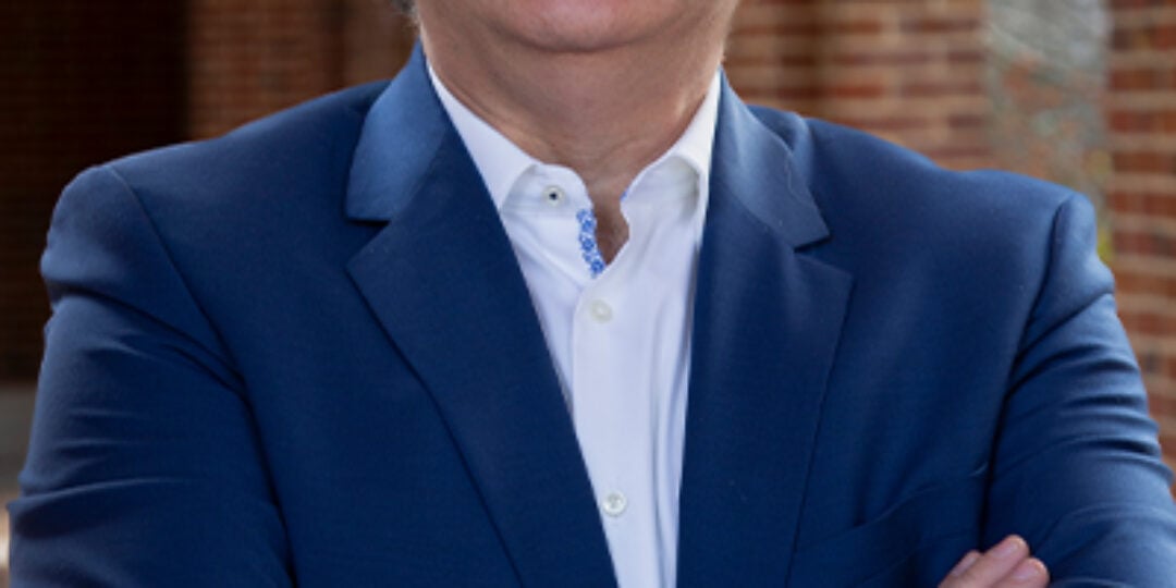 Headshot of Konstantinos Konstantopoulos. He has salt and pepper hair, brown eyes, and light skin tone. He is wearing a dark blue dress jacket and white button down dress shirt. He is standing outside under a brick and marble archway.