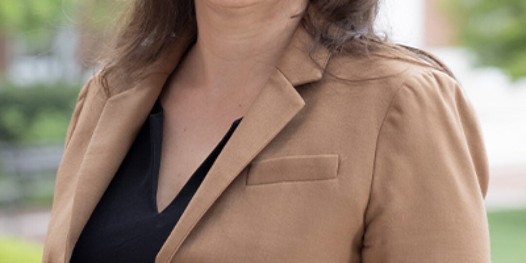 Headshot of Rebecca Schulman. She has long brown hair with bangs, brown eyes, and light skin tone. She is wearing a tan dress jacket over a black v-neck dress shirt. She is standing outside with a red and white brick building and trees in the background.