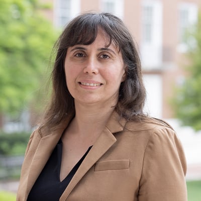 Headshot of Rebecca Schulman. She has long brown hair with bangs, brown eyes, and light skin tone. She is wearing a tan dress jacket over a black v-neck dress shirt. She is standing outside with a red and white brick building and trees in the background.