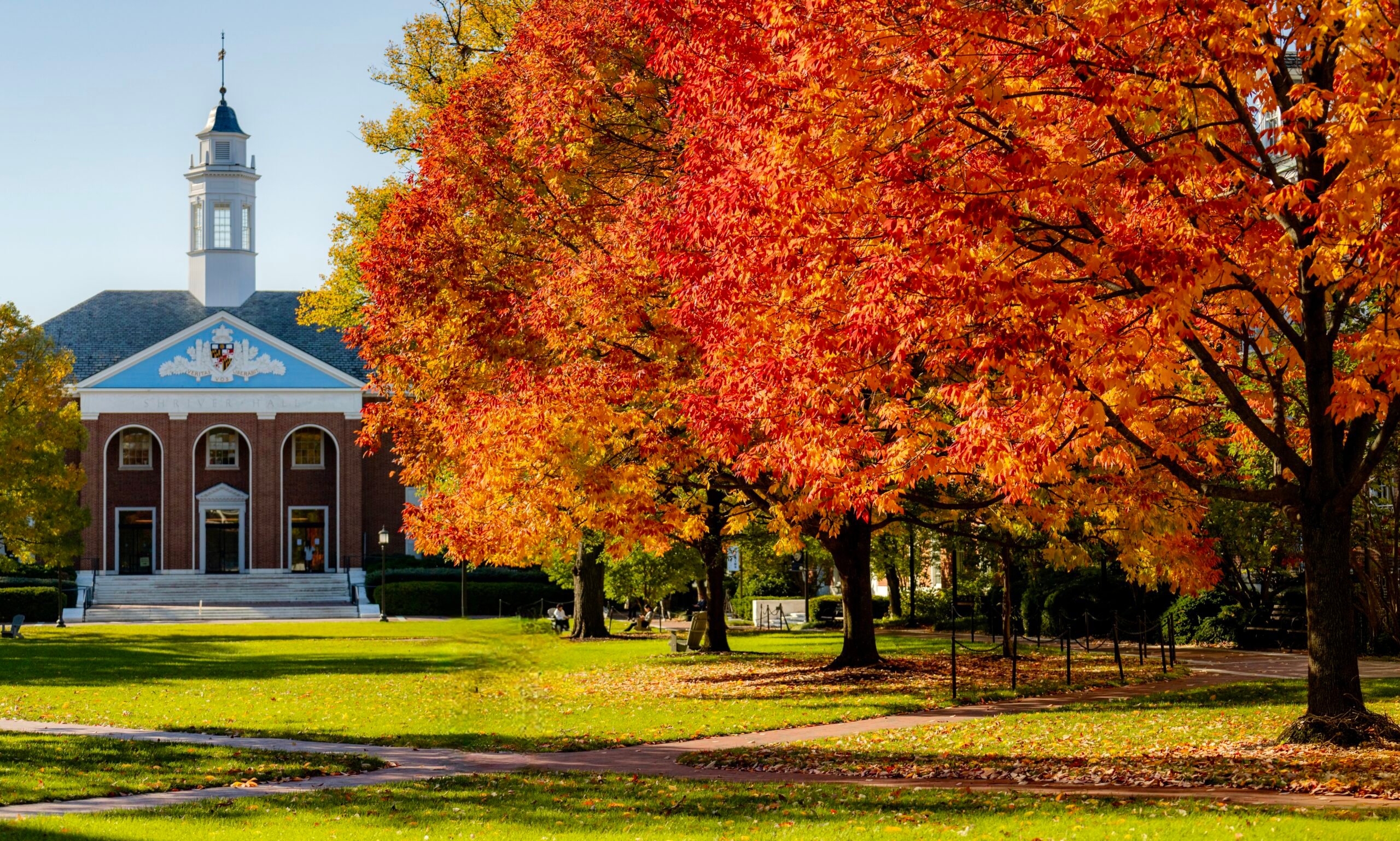 Red and white brick building with white bell tower in the background. On the right are two maple trees with red and orange leaves on the right. In the foreground is an open campus quad with green grass and a brick pathway.