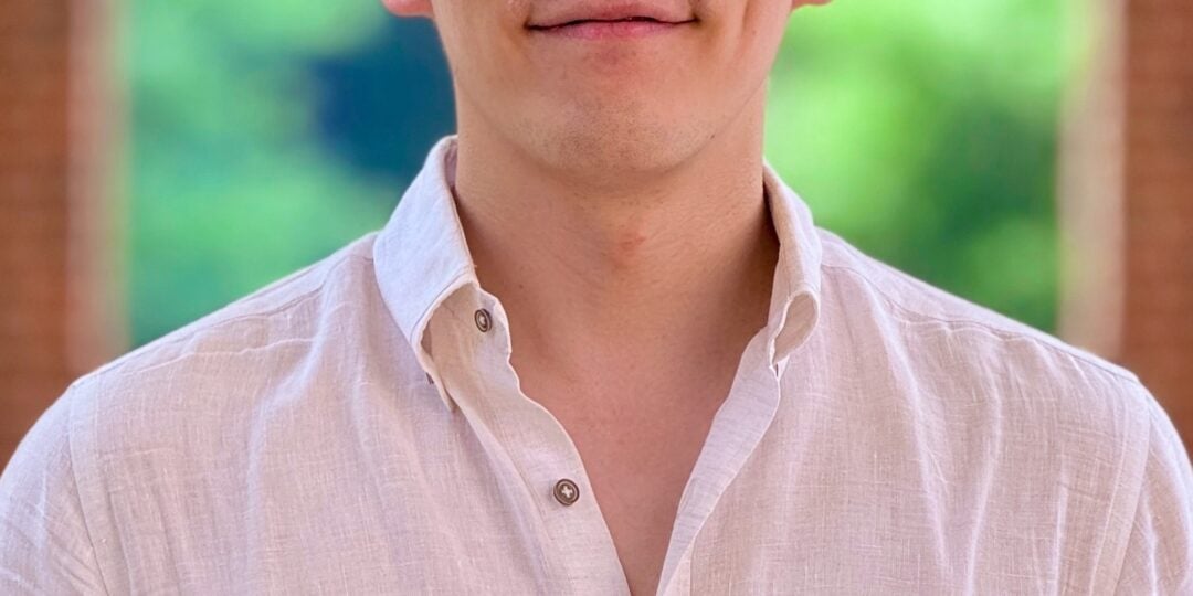 Headshot of Sangkyun Cho. He has short dark hair, brown eyes, and light skin-tone. He is wearing a white button down dress shirt and is standing outside in brick archway with trees in the background.